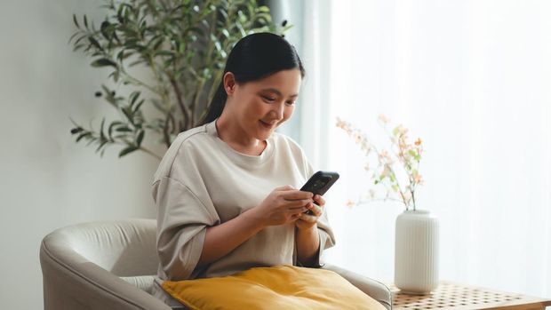 Asian woman sitting on sofa and holding smart phone at home. Happy woman using smart phone for searching shopping online or surfing social media.