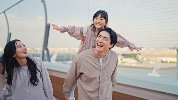 A family is walking outdoors near an airport area. A father is carrying a young child on his shoulders while the child stretches their arms outward. The mother is walking beside them, smiling. Airport infrastructure and aircraft are visible in the background behind a fence. The scene takes place during daytime and shows a family travel moment connected to aviation, transportation, and outdoor movement near an airport.