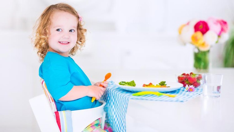 502663557 Happy little girl, cute curly toddler, eating fresh vegetables for lunch, healthy salad snack, corn, broccoli, carrots and strawberry fruit in a white dining room sitting in a high chair