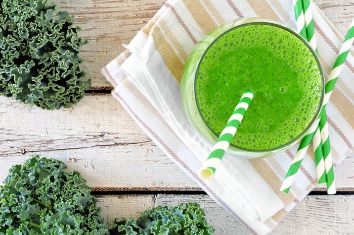 Green kale smoothie overhead view, in a glass with straw on a striped cloth against aged wood