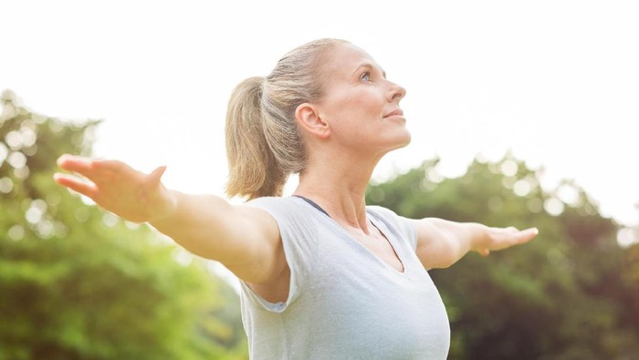 Mature woman doing yoga at park and looking away. Senior blonde woman enjoying nature during a breathing exercise. Portrait of a fitness woman stretching arms and looking away outdoor. 