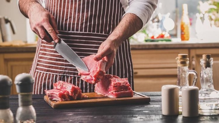Man cuts of fresh piece of meat on a wooden cutting board in the home kitchen. A man in a striped apron with a big knife in his hands