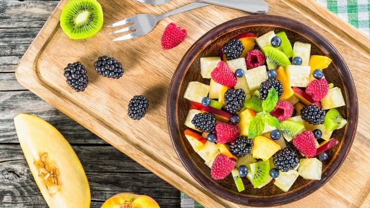 delicious fruit and berry summer salad decorated with mint leaves on clay dish on cutting board with dessert forks, half of peach and slice of melon on rustic boards, view from above