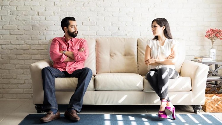 Husband and wife sitting on sofa with their arms crossed and looking at each other after a fight in living room