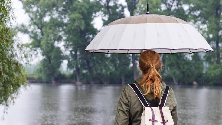 Woman with umbrella and backpack standing in rain at lake