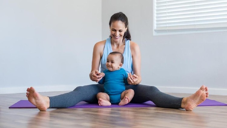 Happy fitness woman doing yoga stretching exercise with infant son. Active mother playing with toddler on floor. Little boy enjoying with mom sitting on purple yoga mat at home.