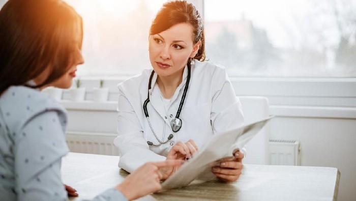 An Asian girl is indoors in a hospital room. She is wearing casual clothing. An Asian female doctor wearing medical clothing is checking her heartbeat with a stethoscope.