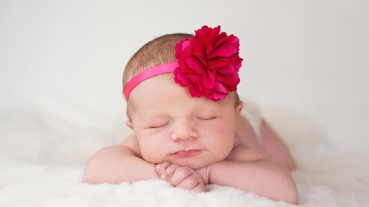 A portrait of a beautiful newborn baby girl wearing a hot pink flower headband. She is sleeping on a cream colored sheepskin rug.