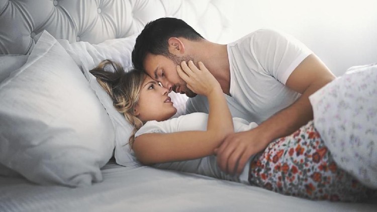 Closeup side view of late 20's couple kissing in bed which probably leads to some love making. They are both still wearing pajamas and everything is decent.