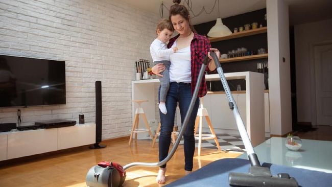 One young woman holding her baby, using vacuum cleaner for cleaninig her house.
