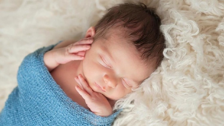 A 12 day old newborn baby boy sleeping on a beige flokati rug and swaddled in a gauzy light blue wrap.