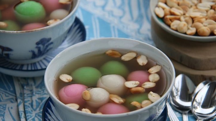 A Chinese Peranakan dessert of glutinous rice balls in warm ginger soup. Served in vintage ceramic bowls with extra peanuts, also in vintage saucer. A few teaspoons are placed on a folded batik napkin. The meal is arranged on a rustic wooden table.