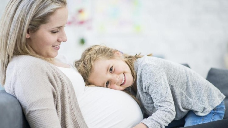hamil A pregnant Caucasian mother and her daughter are indoors in a living room. They are wearing casual clothing. The daughter is laying her head on her mother's stomach.