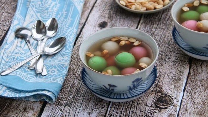 A Chinese Peranakan dessert of glutinous rice balls in warm ginger soup. Served in vintage ceramic bowls with extra peanuts, also in vintage saucer. A few teaspoons are placed on a folded batik napkin. The meal is arranged on a rustic wooden table.