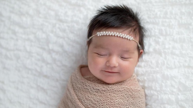 917059906 Smiling one month old baby girl swaddled in a beige wrap. Shot in the studio on a white blanket.