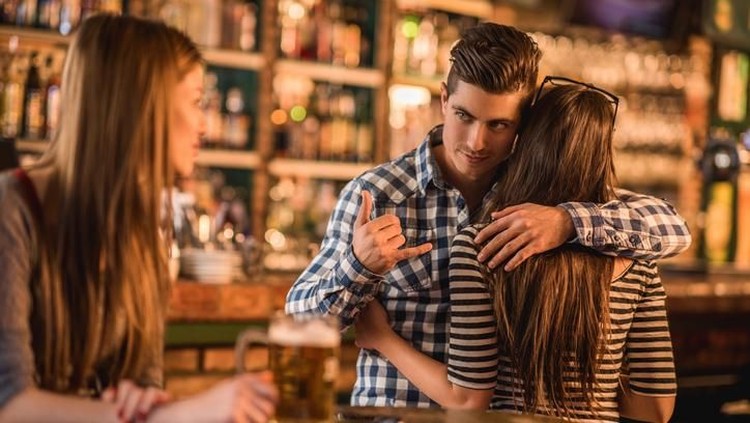 Unfaithful young man in a cafe. He is embracing his girlfriend and making call gesture to another woman behind her back.