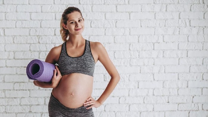 Beautiful pregnant woman workout. Doing fitness on last months of pregnancy. Yoga. Standing on white brick background with sports mat.