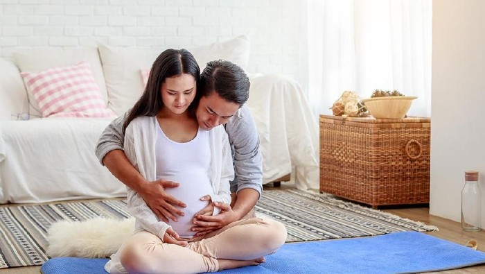 Happy pregnant woman with her husband are holding and listening to baby in belly and setting on living room's floor at home, pregnant and family concept