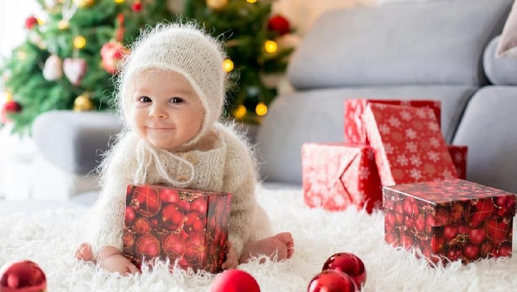 Little baby boy in white knitted onesie, playing with and opening presents at home on Christmas