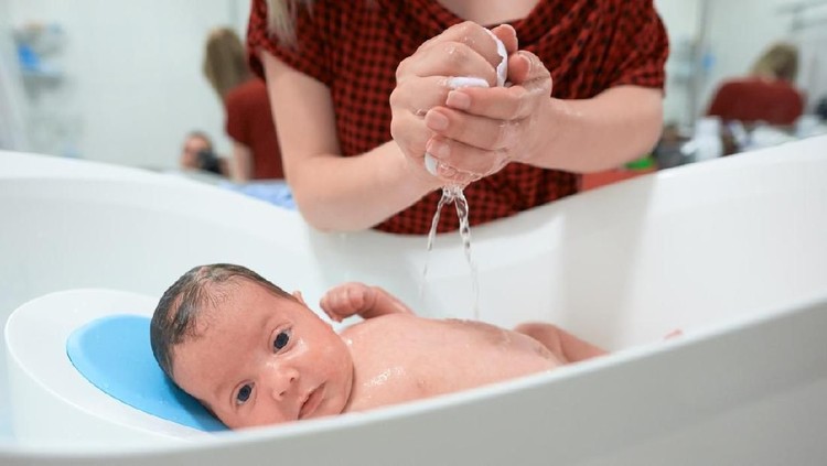 cute baby girl inside bathtub enjoying bath, unrecognizable mother hands washing her.