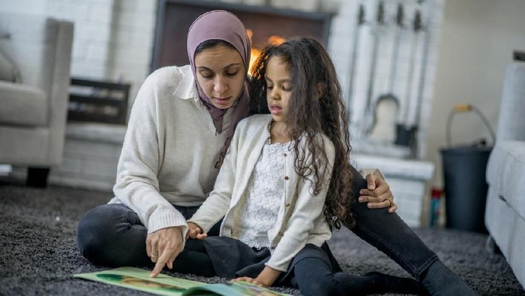 A Muslim mother and her daughter are indoors in their living room. The mother is wearing a head scarf. They are sitting on the carpet and reading a storybook together.