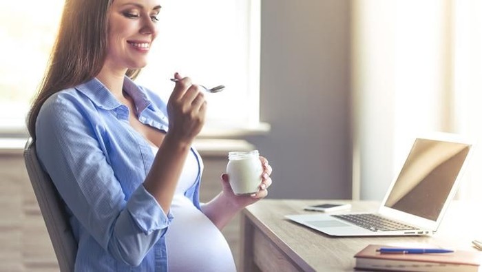 Beautiful pregnant business woman is eating yogurt and smiling while sitting at her working place at home