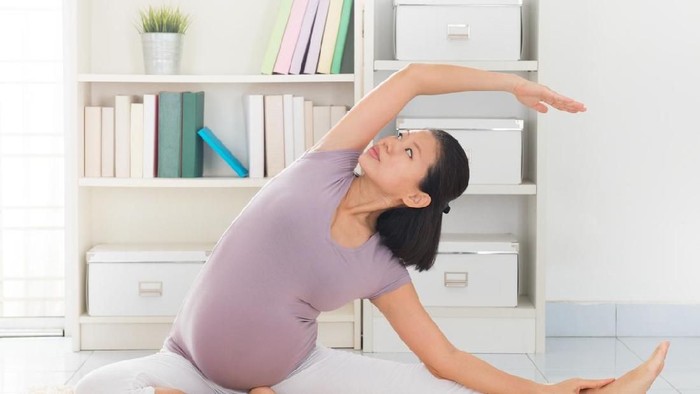 Pregnancy yoga meditation. Full length healthy 8 months pregnant calm Asian woman meditating or doing yoga exercise at home. Relaxation yoga sitting side stretch positions.