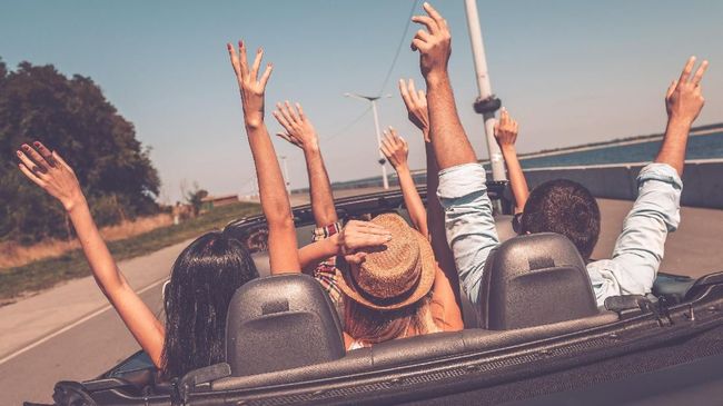 Rear view of young happy people enjoying road trip in their convertible and raising their arms up