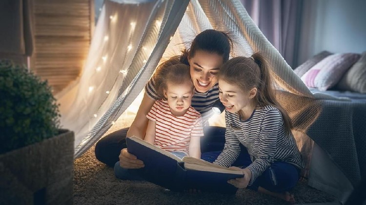 Family bedtime. Mom and children daughters are reading a book in tent. Pretty young mother and lovely girls having fun in children room.