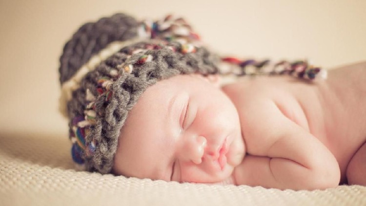 Front view of the face of a cute naked white Caucasian new-born baby boy sleeping on a plain white knitted blanket, hands under the face, wearing a hand knitted hat. Shot on Canon EOS into a studio. Warm blurred sepia edit to emphasize overall softness feel.