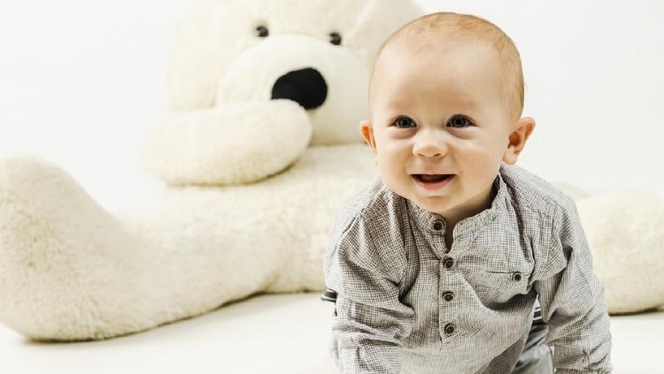 11 months old smiling baby boy in gray clothes is crawling on the white floor against huge teddy bear, studio shot.
