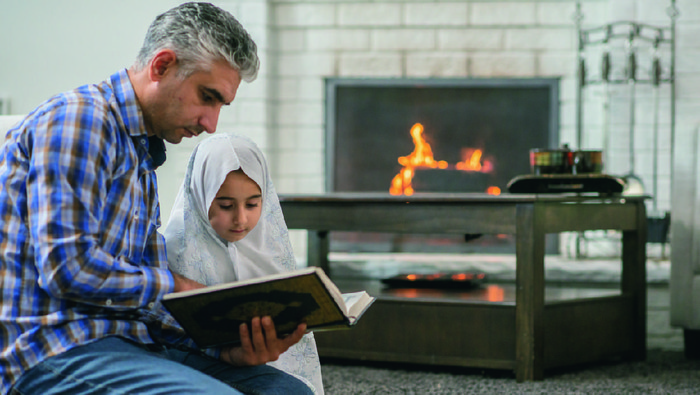 A Muslim father and daughter are indoors in their living room. They are kneeling on the floor to pray. The girl is wearing a traditional shawl.