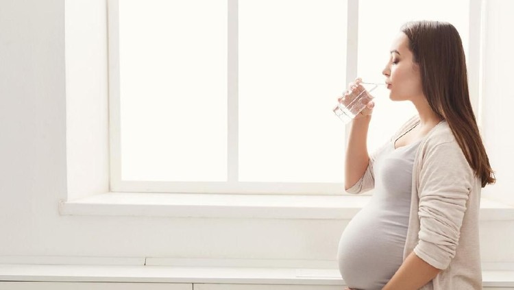 Pensive pregnant woman drinking water near window. Young happy expectant thinking about her baby and enjoying her future life. Motherhood, pregnancy, happiness concept