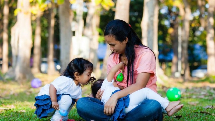 menyusui Breastfeeding. Young mother feeding her baby in the park and other daughter was playing with her sister.