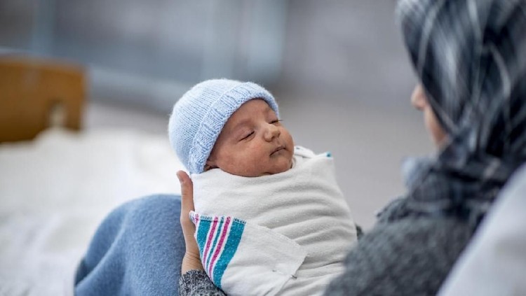 1134381042 A Muslim woman is laying in a hospital bed. She is holding her newborn son in her arms. She is watching over her baby while he sleeps.