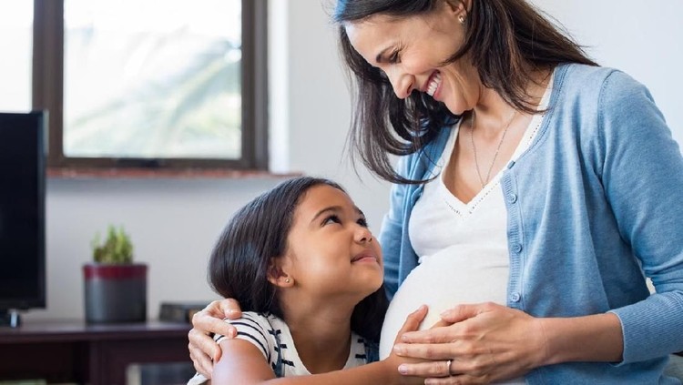 Lovely daughter touching mother's pregnant belly. Happy little girl feeling baby at mother tummy and awaiting the birth of her little brother. Pregnant mother relaxing on sofa with her cute daughter.
