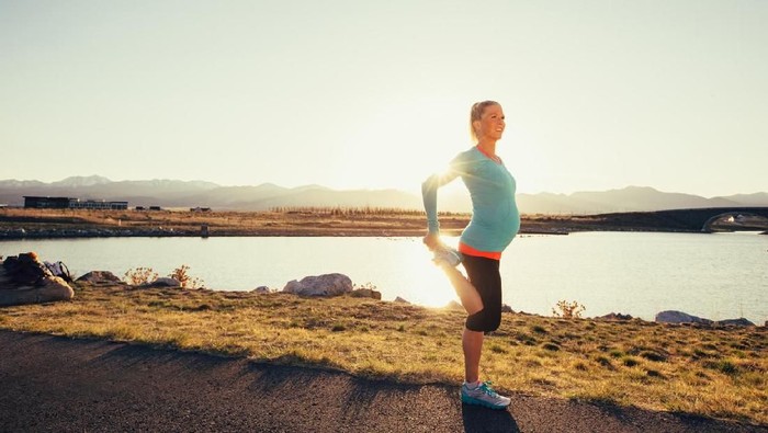 A pregnant female runner in a blue athletic shirt and running shoes is training while pregnant for her next race. She warms up by stretching with the sun flaring behind her. This woman enjoys running on trails and at sunset in the evening. Running is her favorite exercise to stay fit and happy.
