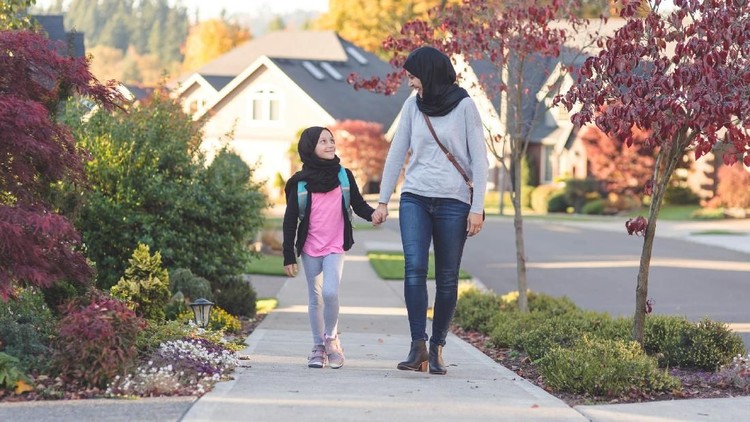 A mother wearing a traditional headscarf picks up and hugs her elementary age daughter, who is also wearing a headscarf,  before she heads off to school. They're on a sidewalk in a suburban neighborhood.
