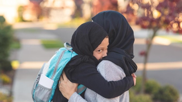 A mother wearing a traditional headscarf picks up and hugs her elementary age daughter, who is also wearing a headscarf,  before she heads off to school. They're on a sidewalk in a suburban neighborhood.
