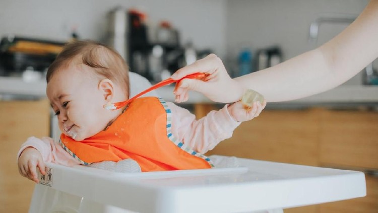 Crying baby boy refusing to eat food from spoon with hands dirty of vegetable puree.