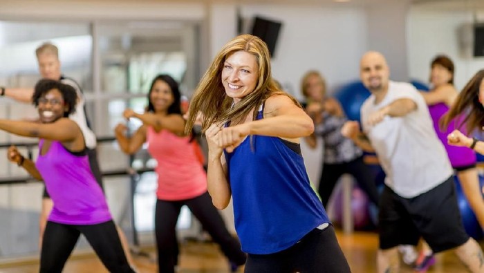 A woman is leading a multi-ethnic boxing class, everyone is wearing athletic clothing and is throwing a punch together - they are smiling and looking at the camera.