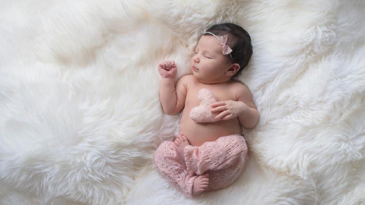 Sleeping, week old newborn baby girl wearing light pink, knitted pants and holding a tiny, heart shaped pillow. Shot in the studio on a white sheepskin rug.