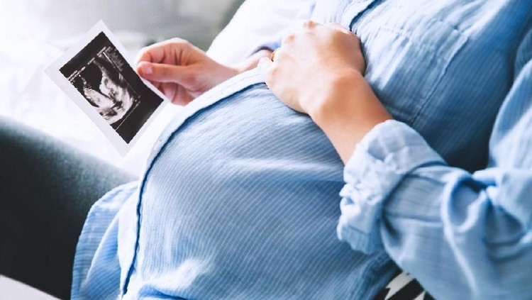usia hamil Pregnant woman keeping hand on belly and holding ultrasound image at home interiors. Pregnancy, parenthood, preparation and expectation concept. Close-up, copy space, indoors.