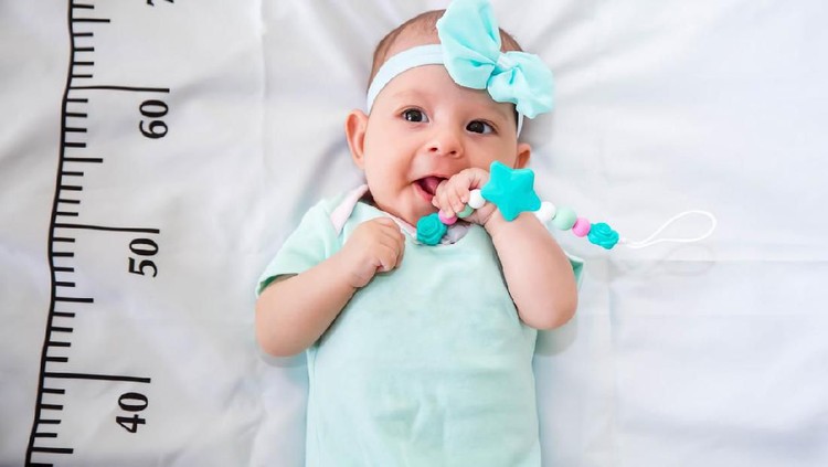 A three month baby in mint green clothes lying on a bed on which a measuring ruler for growth is drawn. Teething tool in hands, chewing