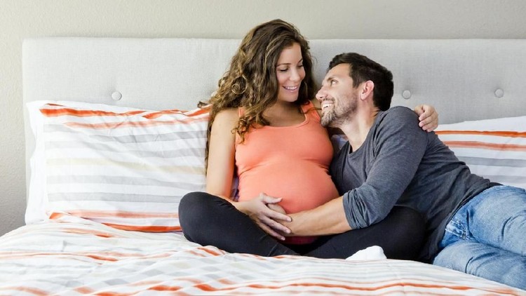 Young happy pregnant couple sitting on a bed and smiling. Man looking at woman. Lifestyle maternity photo.