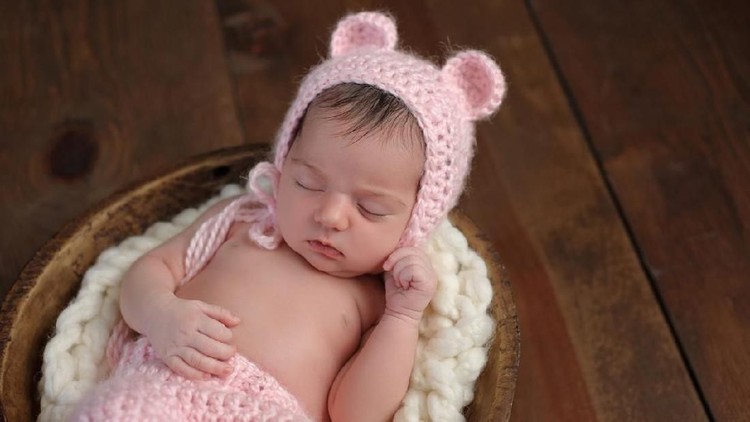 537729274 An overhead shot of a two week old newborn baby girl sleeping in a little, wooden bowl. She is wearing a crocheted, pink bear bonnet. Shot in the studio on a wood background.