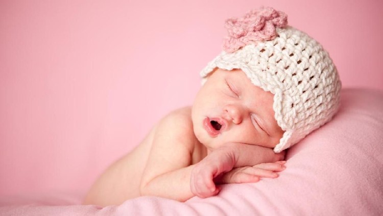 An overhead shot of a two week old newborn baby girl sleeping in a little, wooden bowl. She is wearing a crocheted, pink bear bonnet. Shot in the studio on a wood background.