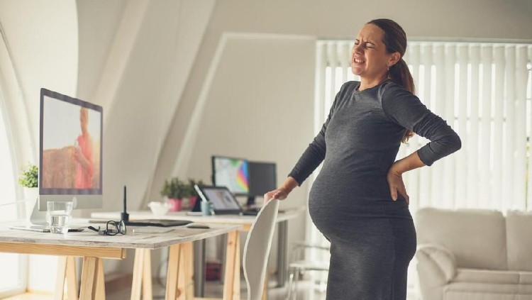 619062186 Pregnant mid adult woman standing by the desk in the office, having back pain at the end of pregnancy.