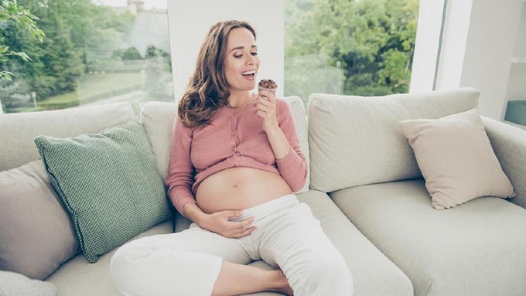 Lovely nice beautiful adorable funny cheerful positive pregnant curly-haired mum in casual wear sitting on sofa, couch, divan, eating yummy cake, white light interior room