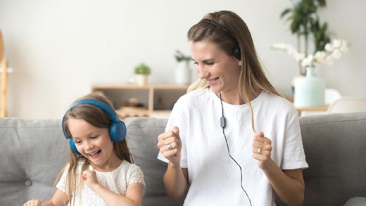 Happy young mother and funny child daughter having fun laughing dancing sitting on couch listening to music in earphones together, smiling mom and kid wearing headphones enjoy favorite songs at home